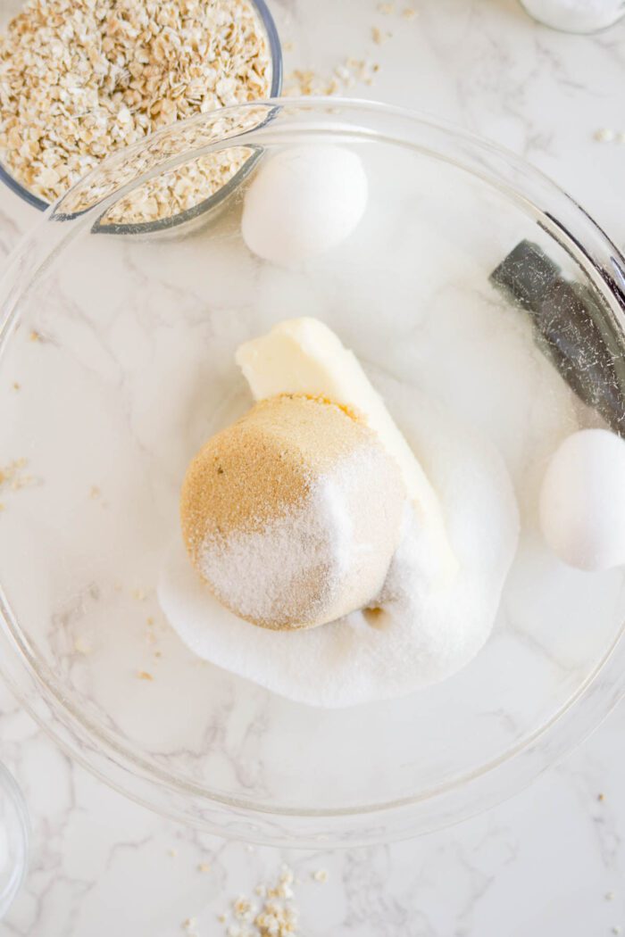 Ingredients for baking the best oatmeal raisin cookies in a glass bowl: brown sugar, white sugar, butter, and eggs, with oats and raisins scattered on a marble surface.