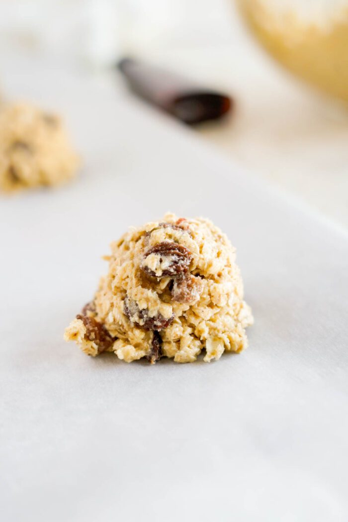 Scoop of raw oatmeal raisin cookie dough on parchment paper, with a mixing bowl and spoon blurred in the background.
