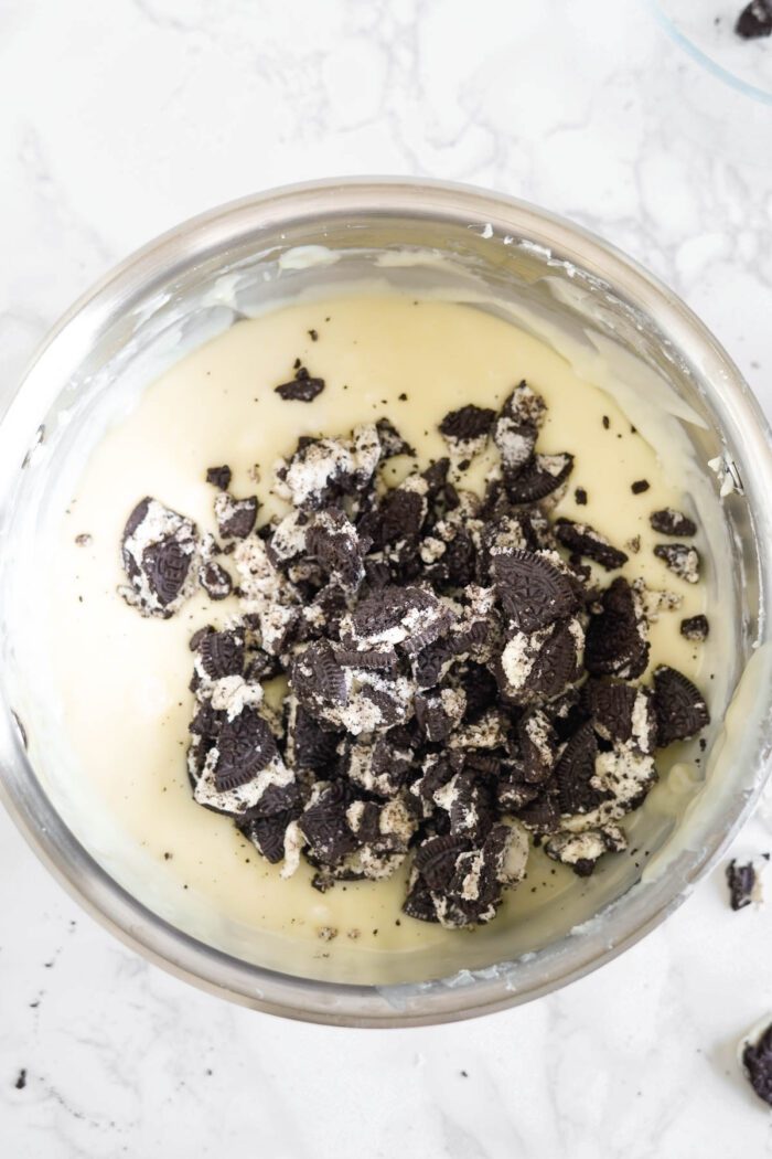 Crushed cookies mixed into a bowl of fudge batter on a marble surface.