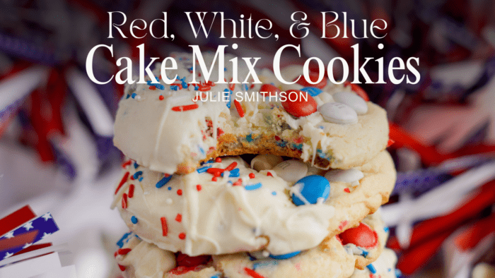 A close-up of a stack of red, white, and blue cake mix cookies with colorful sprinkles and candies, set against a backdrop of festive ribbons. Text overlay reads, "Easy 4th of July Cake Mix Cookies - Julie Smithson.