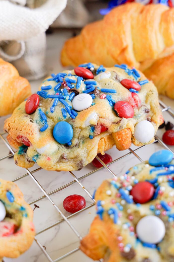 Colorful cookies topped with blue, red, and white candy pieces and sprinkles are placed on a cooling rack, accompanied by freshly baked croissants in the background—perfect for adding to your chocolate chip cookie croissant recipe.