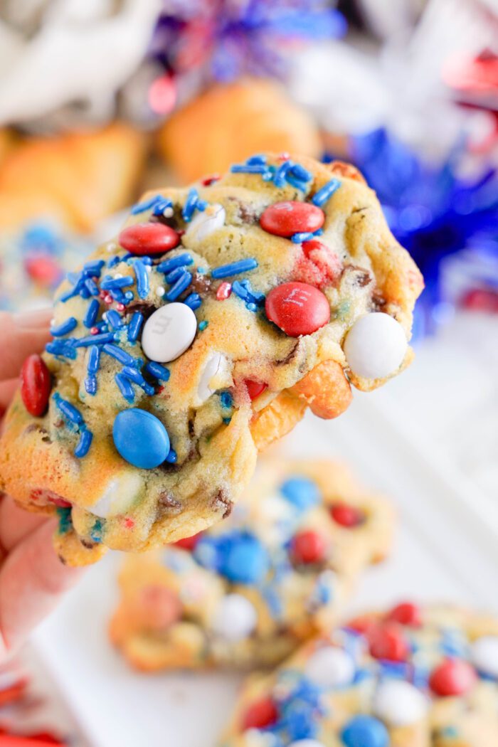 Close-up of a hand holding a cookie topped with red, white, and blue candies and sprinkles. More similarly decorated cookies are visible in the background, reminiscent of a festive twist on a classic chocolate chip cookie croissant recipe.