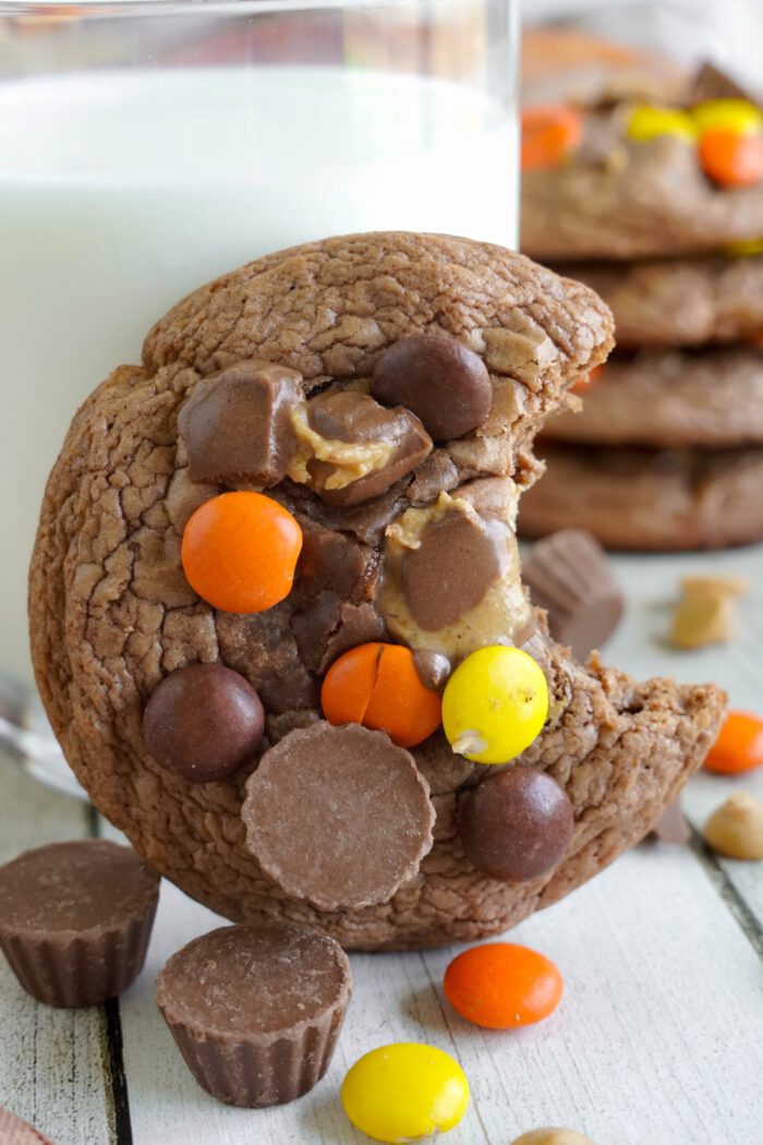 A stack of chocolate chip cookies with reese's pieces and mini peanut butter cups on a wooden table, accompanied by a glass of milk.