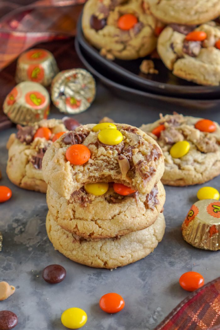 Stack of chocolate chip cookies with colorful candy pieces on a metal surface, surrounded by more candies and cookies.