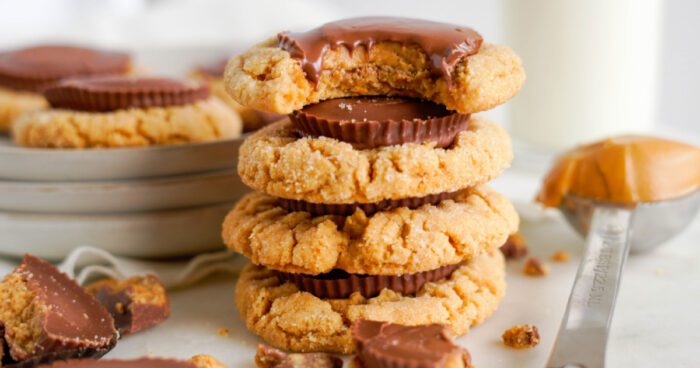 A stack of peanut butter cookies with a chocolate-covered peanut butter cup in the middle, next to broken chocolate pieces and a spoon of peanut butter.