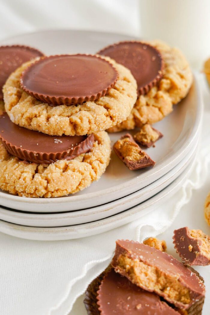 A stack of peanut butter cookies topped with chocolate peanut butter cups on a white plate, with crumbs scattered around.