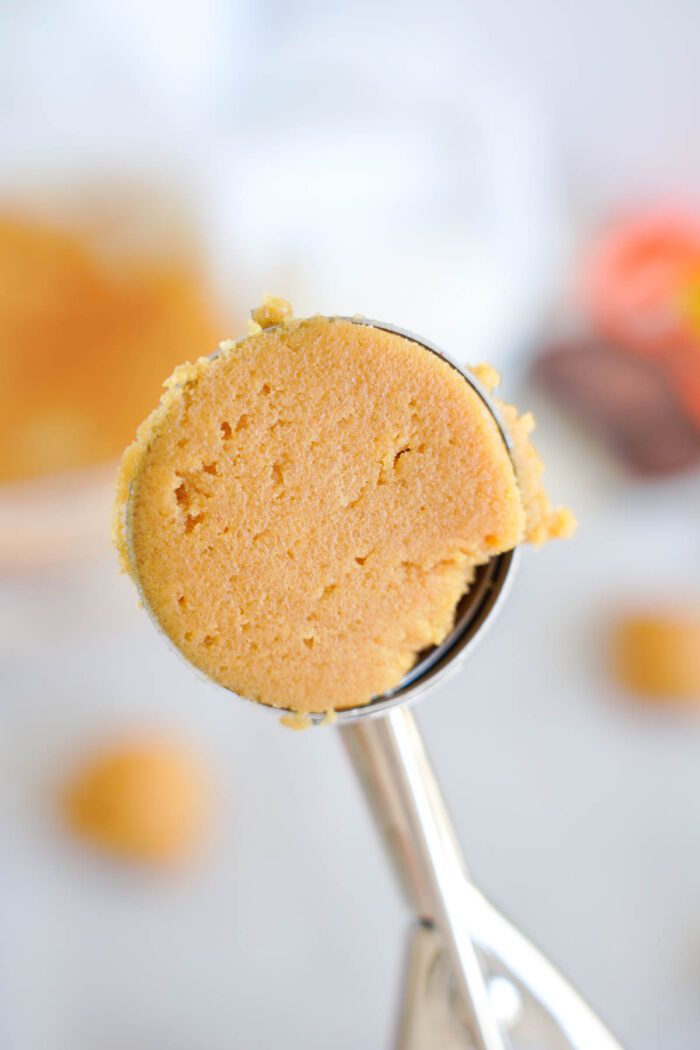 A close-up of a metal ice cream scoop holding a ball of cookie dough for Easy Peanut Butter Cup Cookies, with a blurred background emphasizing additional cookie dough portions.