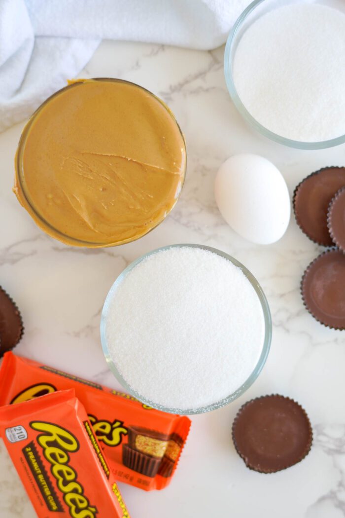 Ingredients for an easy peanut butter cup cookies recipe including peanut butter, sugar, an egg, and Reese's cups on a kitchen counter.