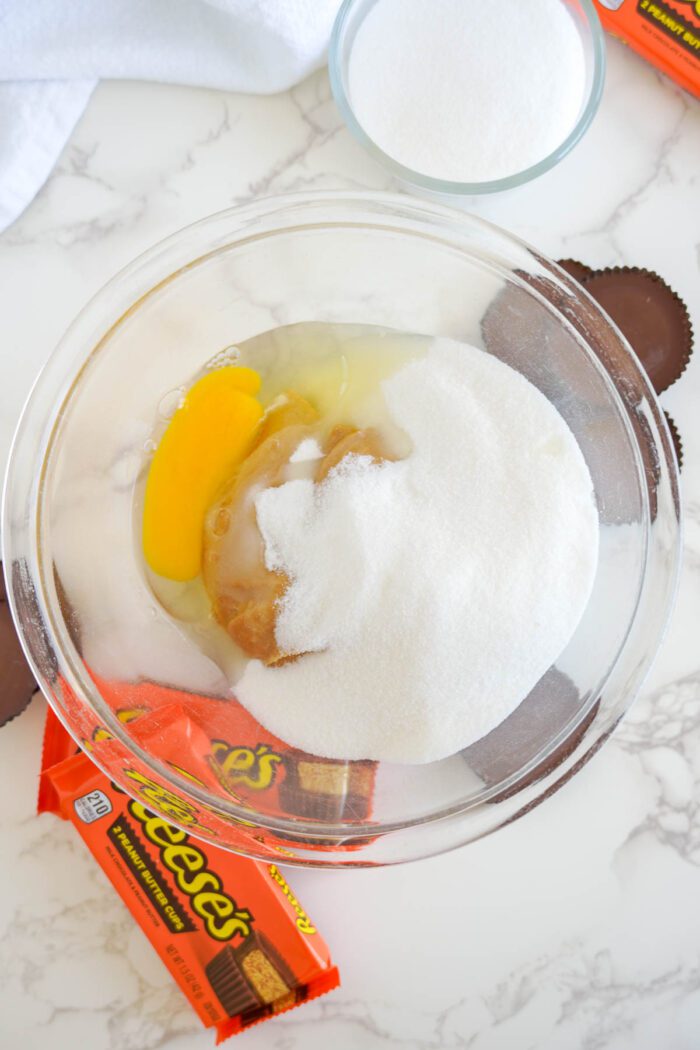 A glass bowl containing eggs, sugar, and a candy wrapper on a marble countertop with a towel and cup of sugar nearby for an Easy Peanut Butter Cup Cookies recipe.