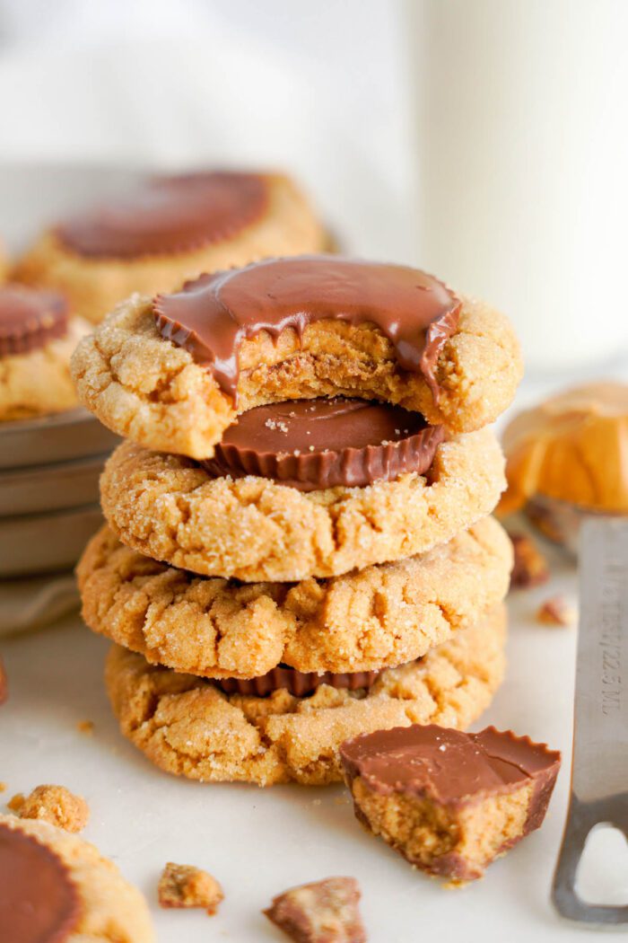 Stack of easy peanut butter cup cookies with a chocolate candy piece on top, beside a glass of milk and scattered candy pieces.
