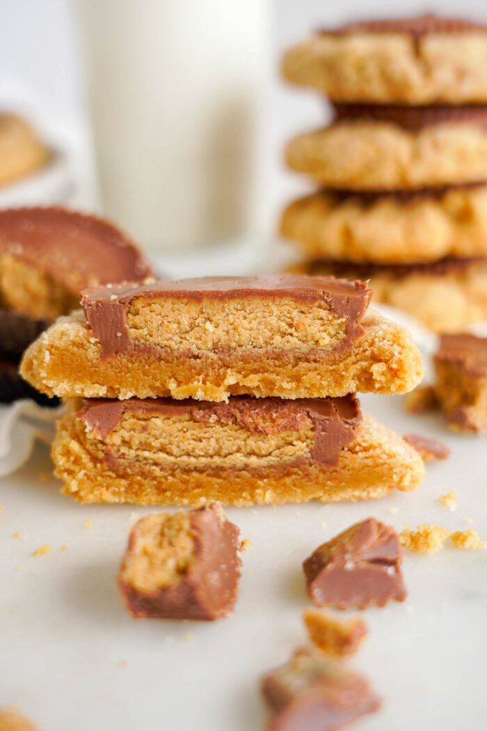 Stack of peanut butter cup cookies with a thick layer of chocolate filling, half a cookie in the foreground showing a cross-section, and crumbs scattered around on a white surface.