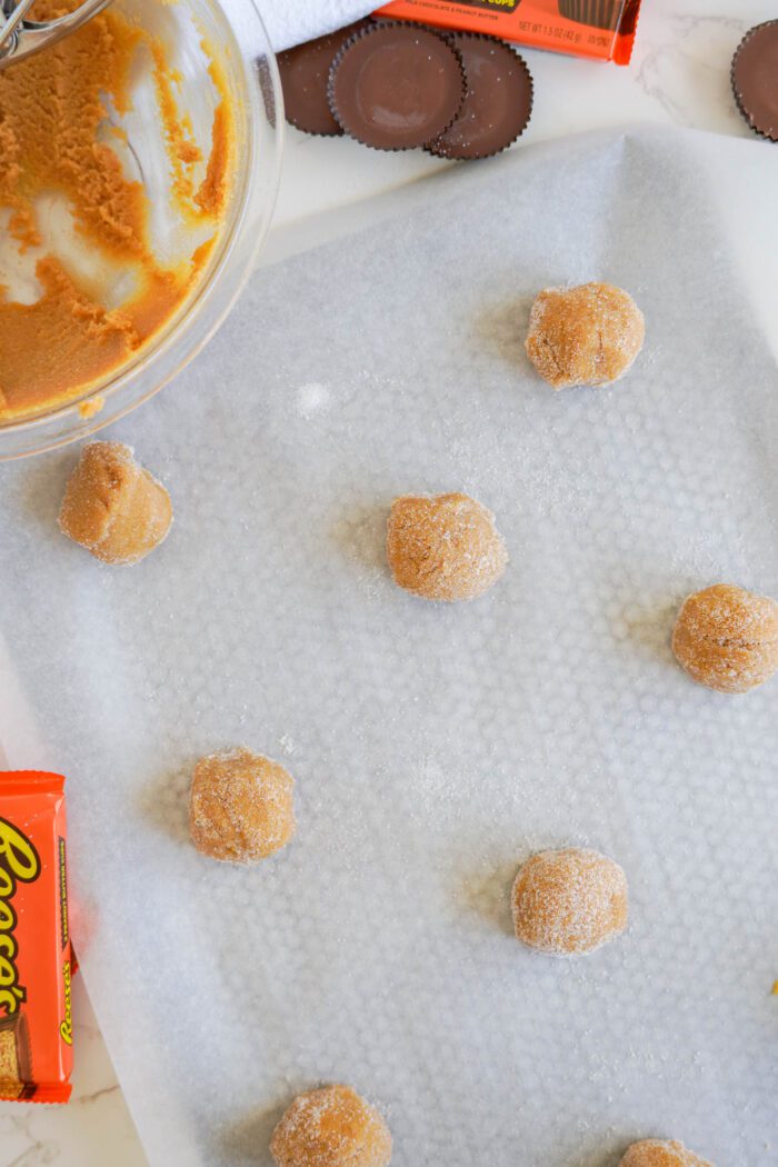 Dough balls for Easy Peanut Butter Cup Cookies Recipe coated in sugar placed evenly on parchment paper, next to a mixing bowl and Reese's cups packs.
