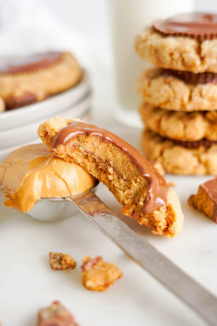 A spoon smearing peanut butter on a cookie with a chocolate piece on top, surrounded by more cookies on a white surface for an easy recipe.