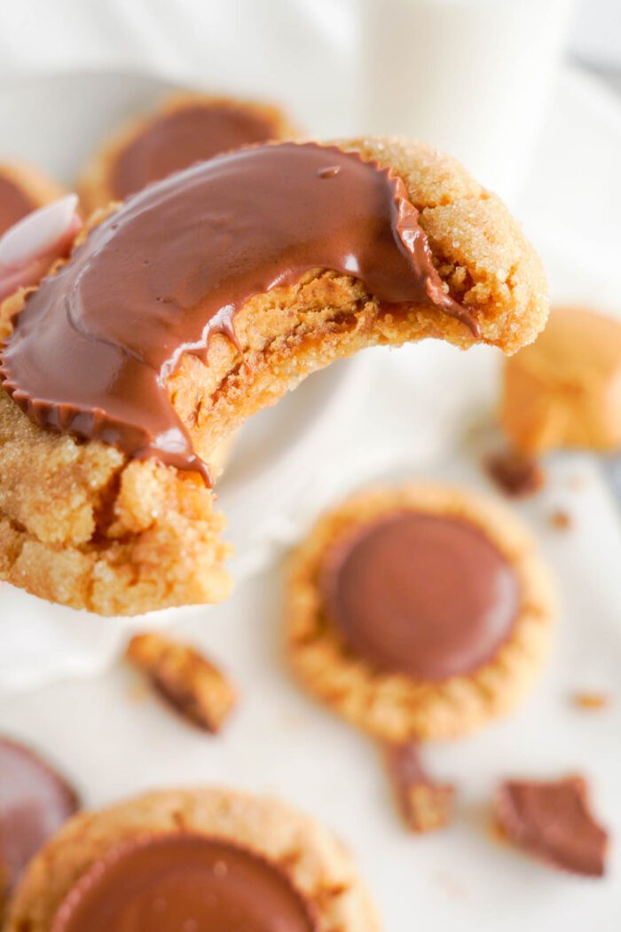 A close-up of an easy peanut butter cup cookie with a chocolate piece, partially bitten, revealing a gooey chocolate filling, with cookie crumbs and chocolate pieces scattered around.