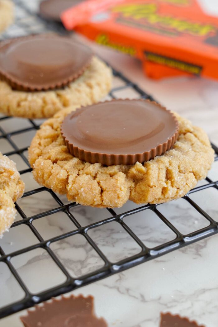 Easy Peanut Butter Cup Cookies Recipe: Peanut butter cookies topped with chocolate peanut butter cups on a cooling rack, with a box of peanut butter cups in the background.