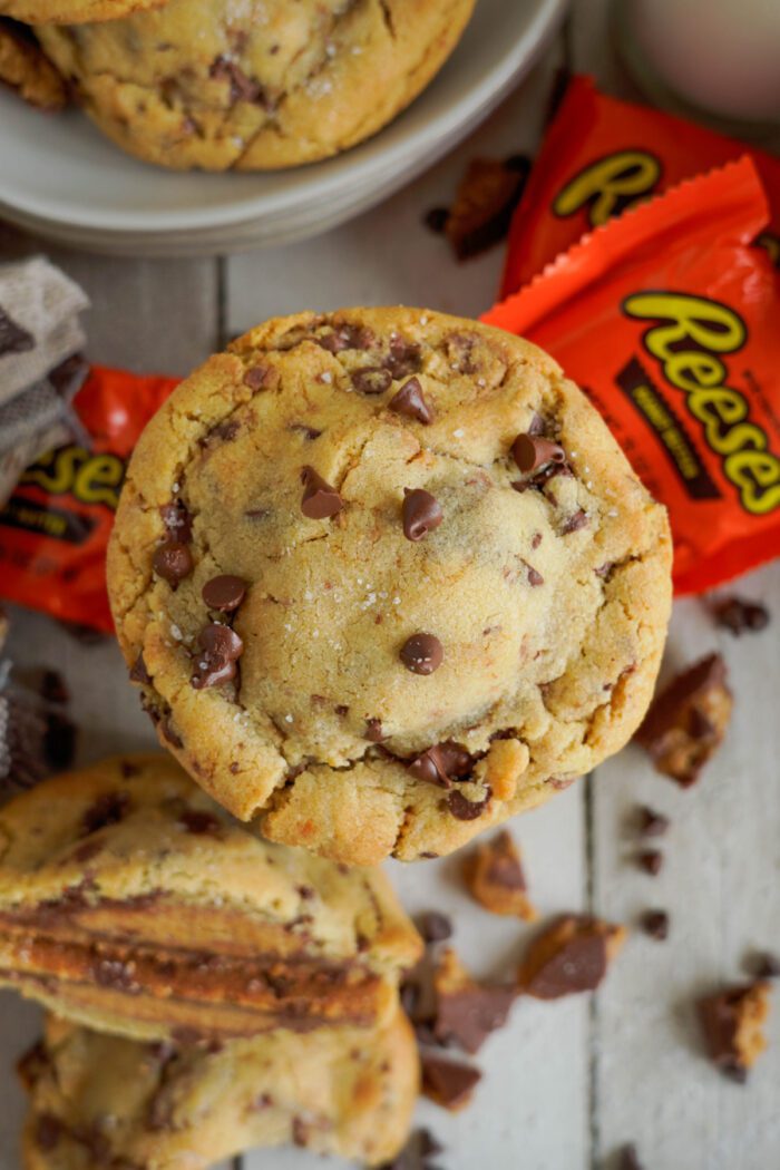 Stack of freshly baked peanut butter cookies with chocolate chips, surrounded by reese's candy wrappers.