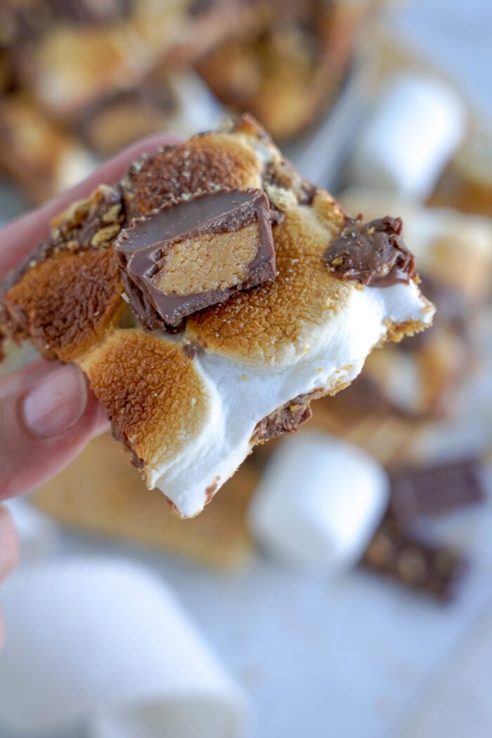 A close-up of a hand holding a s'mores bar with visible layers of graham cracker, melted marshmallow, and chocolate.