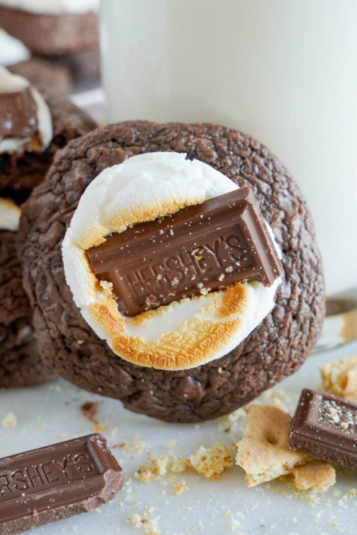 A S'mores Cookie topped with a marshmallow and a piece of Hershey's chocolate, with crumbs and a glass of milk in the background.