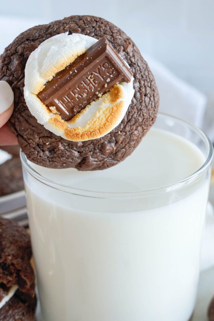 A hand holding a Chocolate S'mores Cookie topped with a toasted marshmallow and a piece of chocolate, above a glass of milk.