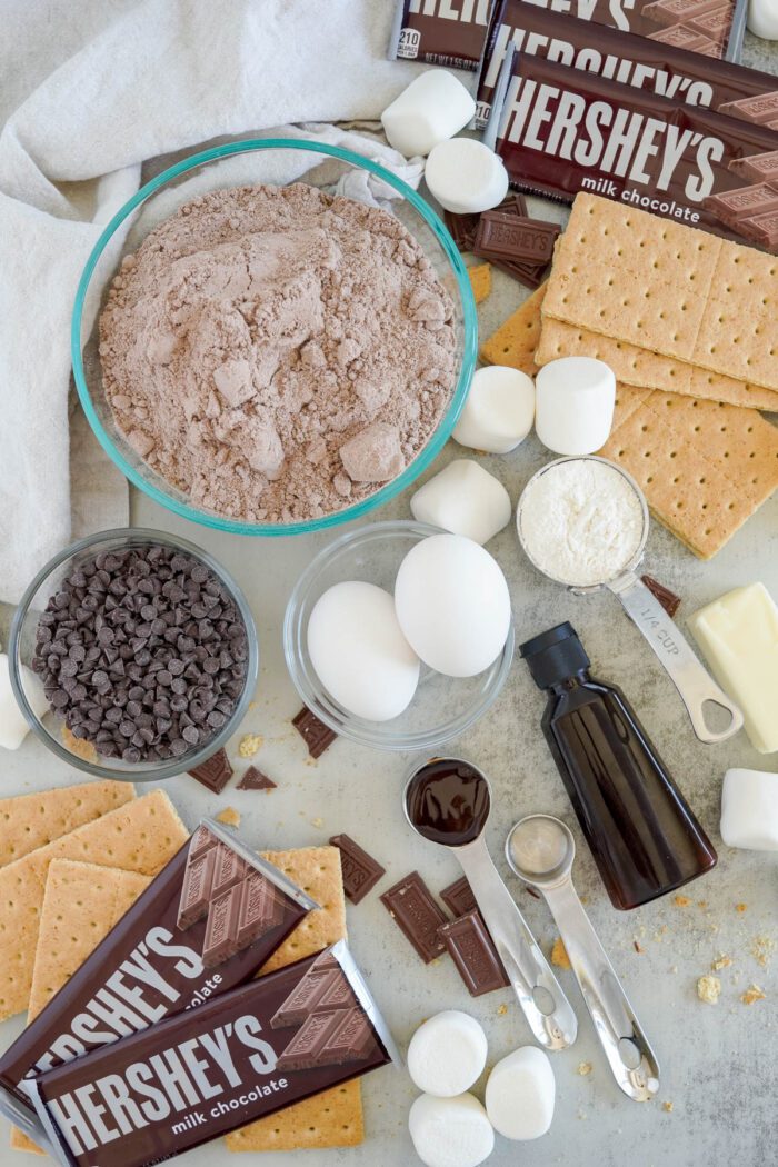 Ingredients for making S'mores Cookies, including graham crackers, marshmallows, milk chocolate bars, and chocolate chips, arranged neatly on a kitchen surface.