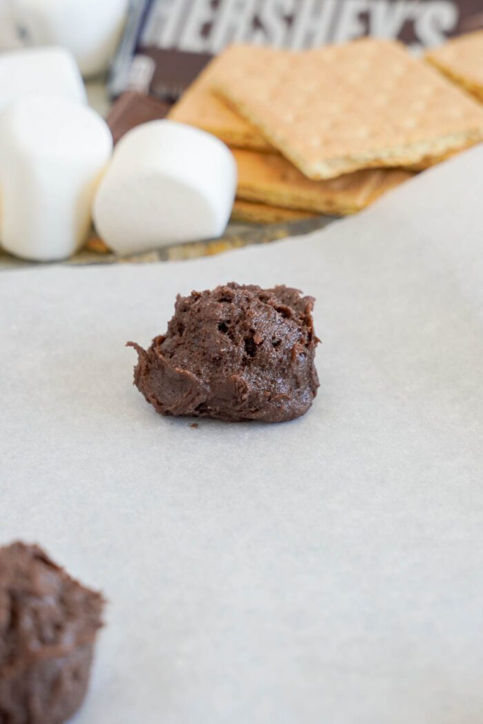 A scoop of S'mores cookie dough balls on parchment paper, with marshmallows, graham crackers, and a Hershey's chocolate bar blurred in the background.