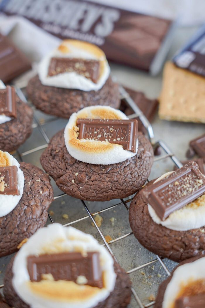 S'mores cookies topped with toasted marshmallows and small pieces of a Hershey's chocolate bar, displayed on a wire cooling rack.