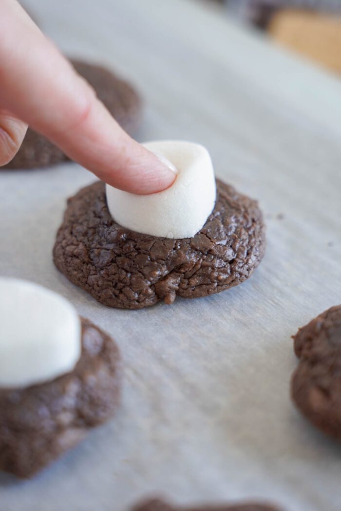 A person pressing a marshmallow onto a freshly baked chocolate cookie on a baking sheet.