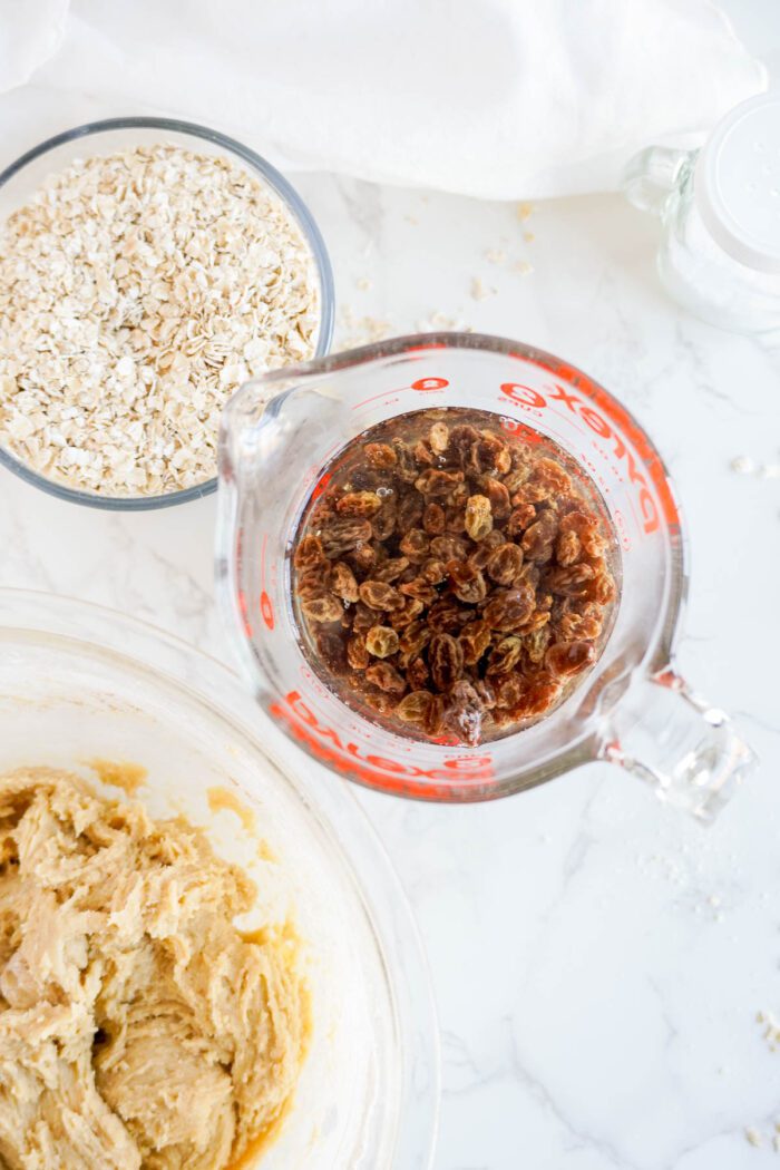 Glass measuring cup filled with raisins beside a bowl of cookie dough for the best oatmeal raisin cookies recipe and a container of oats on a marble countertop.