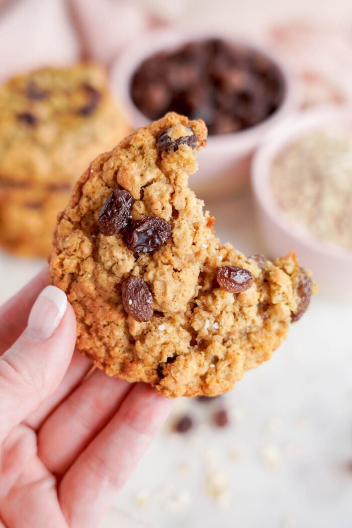 Close-up of a hand holding a half-eaten oatmeal raisin cookie, with more cookies and ingredients for the best oatmeal raisin cookies recipe in the background.