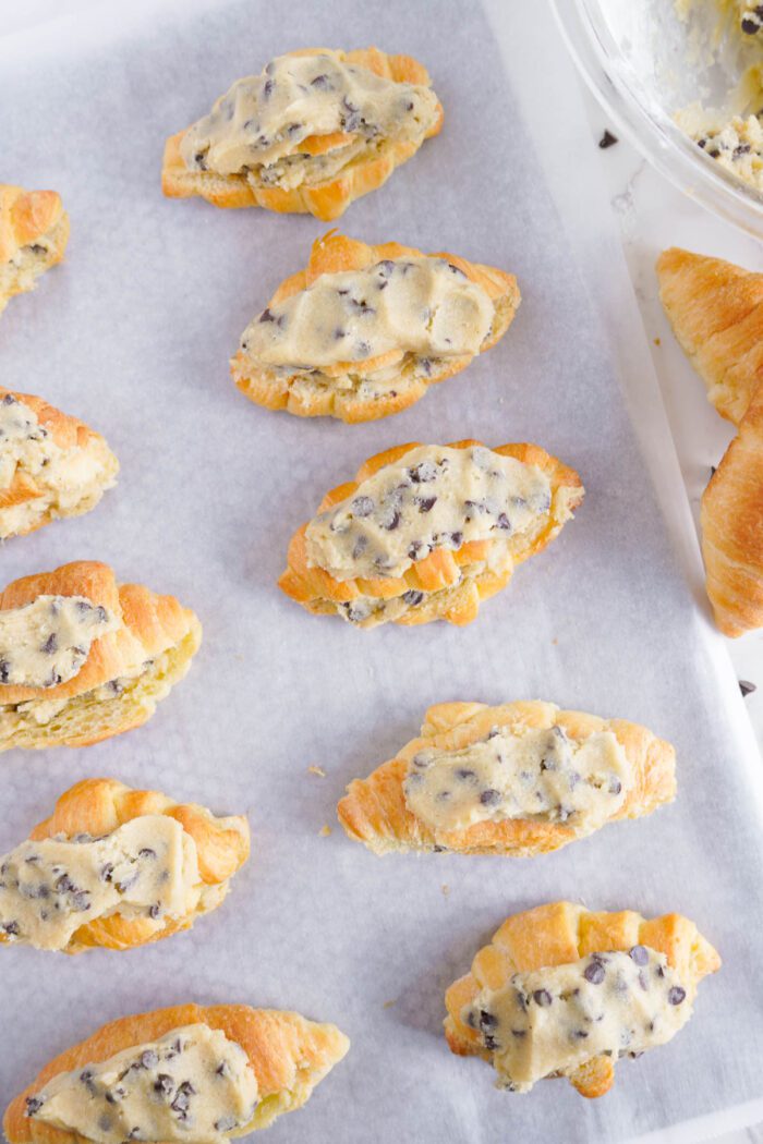 Freshly baked chocolate chip cookie croissants on parchment paper, with a bowl of dough visible in the background.