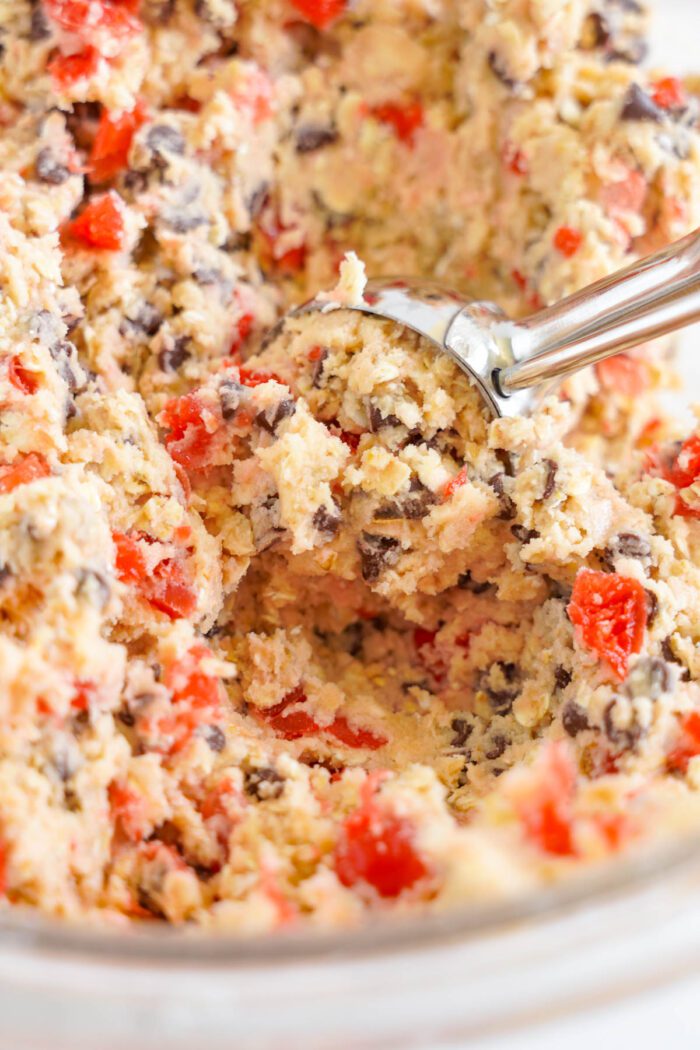 A close-up of Cherry Chocolate Chip Cookie dough with chocolate chips and red cherry pieces, scooped by a metal spoon.