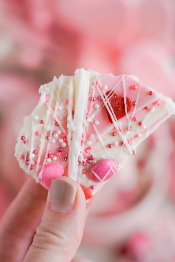 A hand holding a white chocolate bark sprinkled with red and pink candies and sugar beads.
