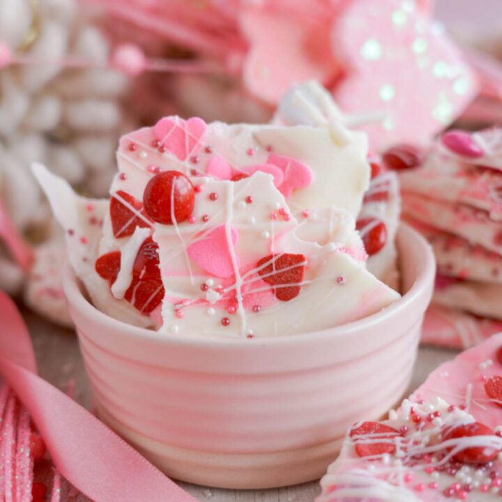 A bowl of white chocolate bark with red candy pieces and sprinkles, surrounded by pink ribbons and more candy bark.