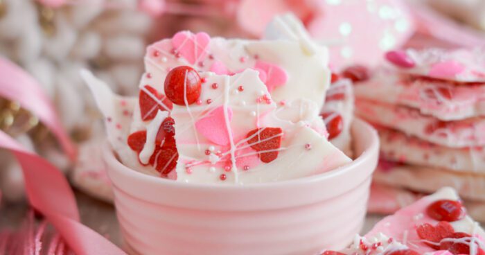 A bowl of Chocolate Candy Bark with pink and red heart-shaped sprinkles and candy pieces, set on a table with thematic decorations.