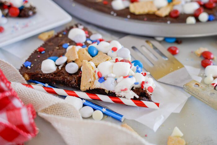 A slice of dessert pizza topped with marshmallows, red, white, and blue candies, and pieces of cookies on wax paper. A red and white striped straw and a silver spatula are partially visible.