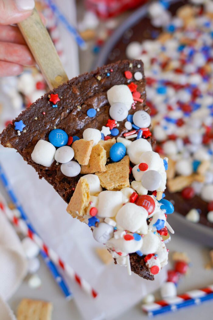 A slice of brownie pizza topped with mini marshmallows, graham cracker pieces, red, white, and blue candies, and festive sprinkles on a wooden stick.