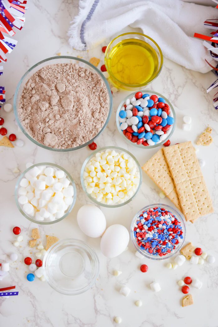 Various baking ingredients including chocolate powder, oil, eggs, water, mini marshmallows, white chocolate chips, M&M's, sprinkles, and graham crackers are arranged on a marble countertop.