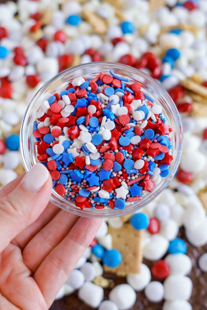 A close-up of a hand holding a small bowl filled with red, white, and blue sprinkles. A mix of marshmallows and sprinkles can be seen in the background.