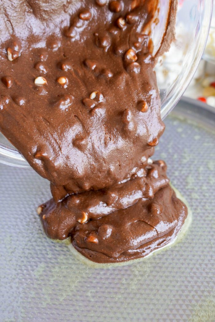 Thick chocolate batter with white chocolate chips is being poured from a bowl onto a greased baking sheet.