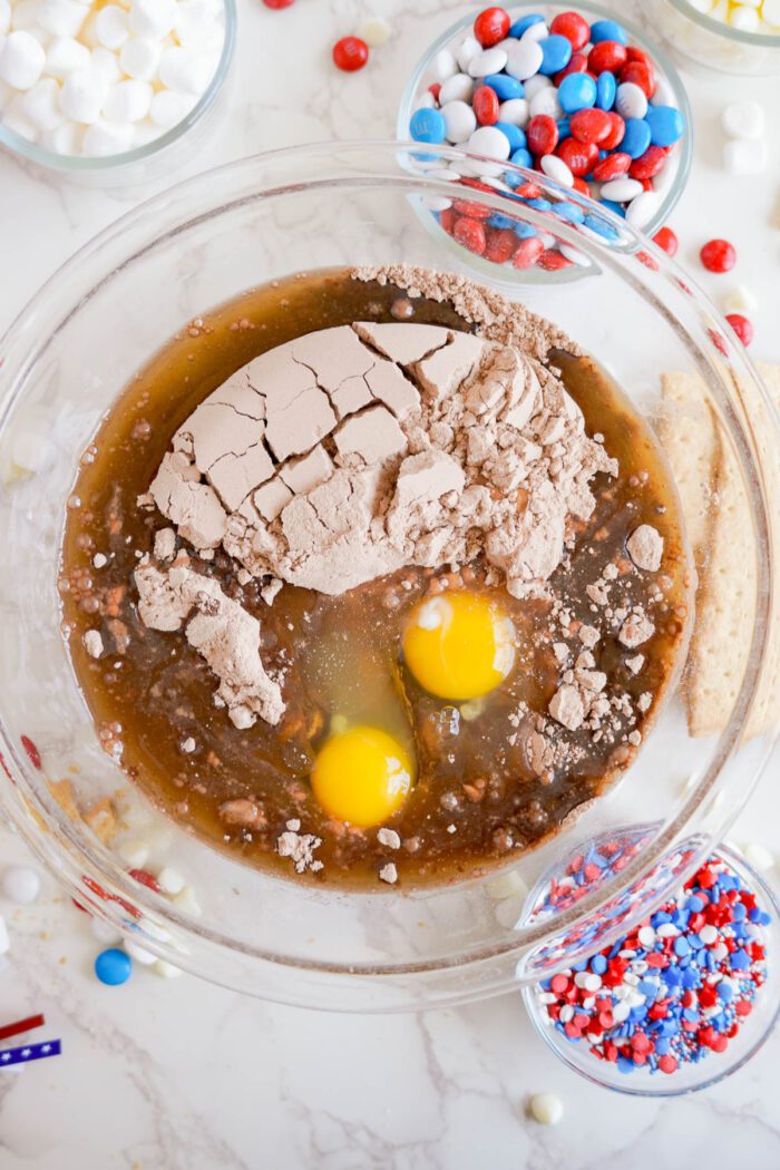 A mixing bowl containing brownie batter with two cracked eggs and dry ingredients. Surrounding the bowl are various small bowls filled with marshmallows and red, white, and blue candies.