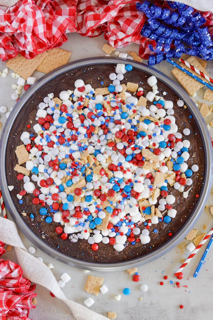 A round brownie is topped with white frosting, red, white, and blue candies, and crushed graham crackers, surrounded by red, white, and blue decorations.