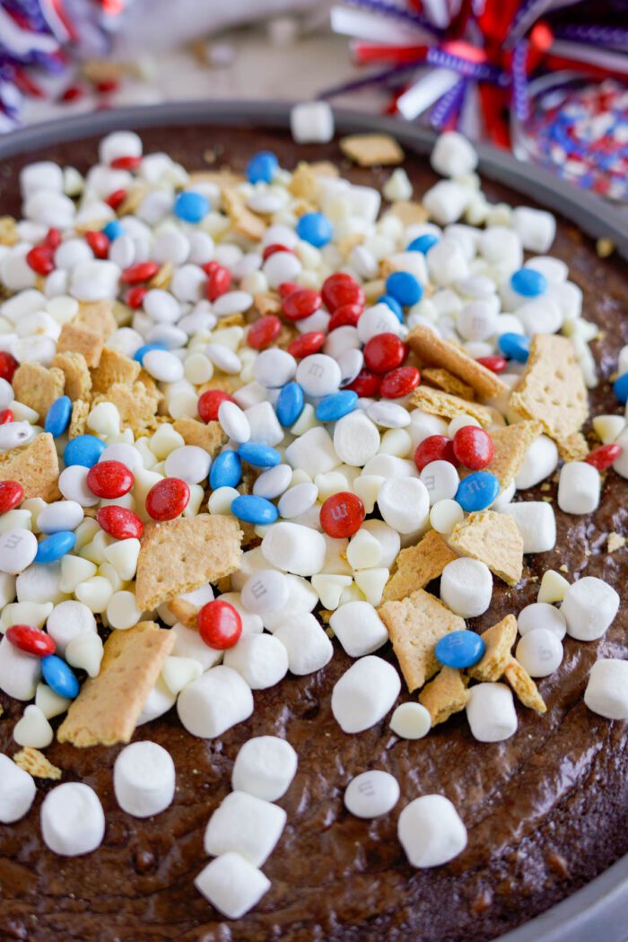 A close-up of a dessert featuring a chocolate base, topped with mini marshmallows, white chocolate chips, broken graham crackers, and red, white, and blue candies.