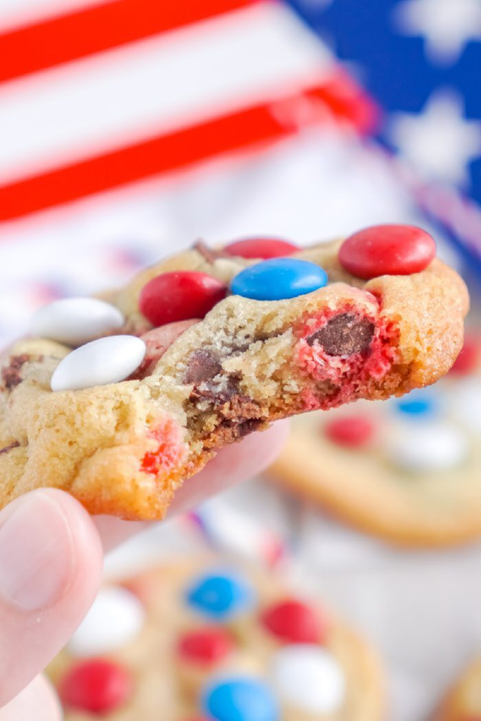 A hand holds a bitten cookie with red, white, and blue candies. An American flag is blurred in the background.