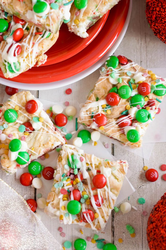 Christmas Cookie bars topped with red and green candy-coated chocolates, white drizzle, and colorful sprinkles, arranged on a wooden surface near a red plate.