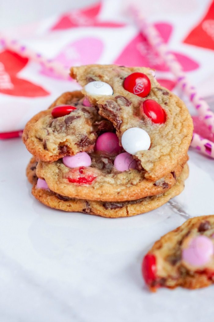 A stack of cookies with red, white, and pink candy pieces on a white surface. One cookie has a bite taken out of it. A pink and red patterned napkin is in the background.