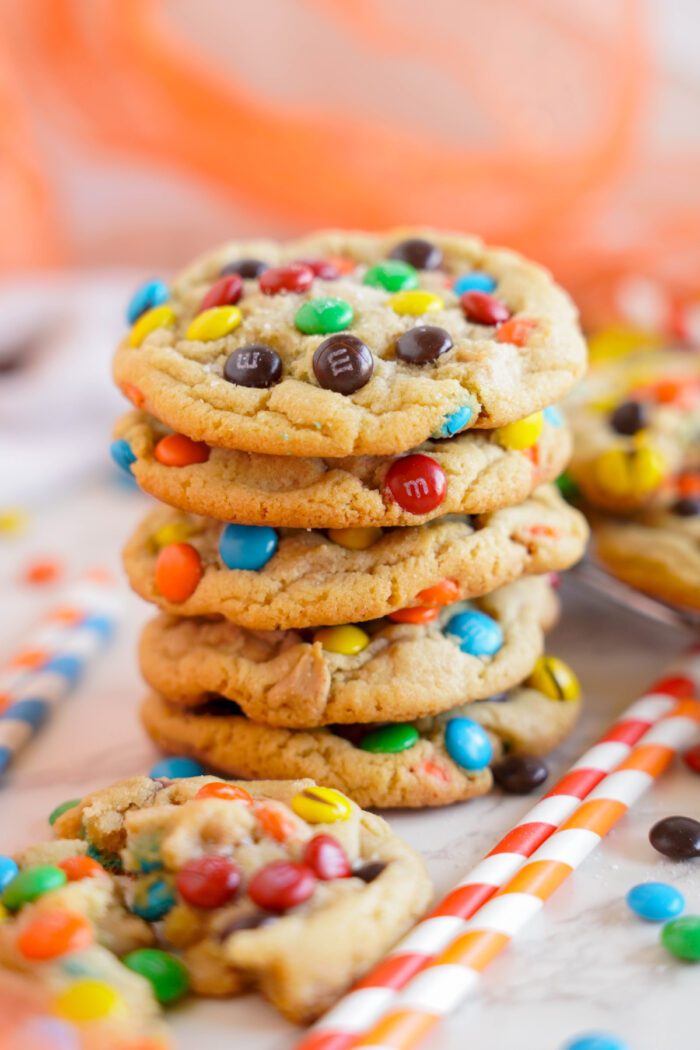 A stack of five cookies with M&Ms on top on a white surface. A broken cookie and striped straws are in the foreground, with an orange backdrop.