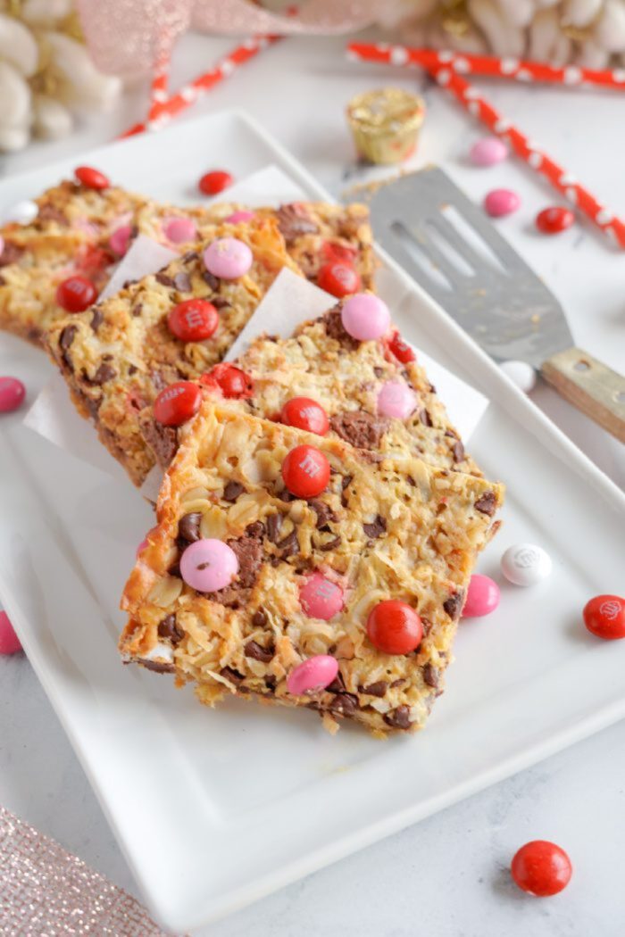 Three dessert bars with chocolate chips, coconut flakes, and colorful candy pieces are arranged on a white rectangular plate. A metal spatula is placed beside them.