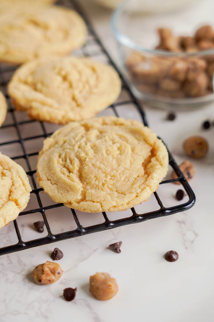 Freshly baked chocolate chip cookies from an easy cookie dough cookies recipe are cooling on a black wire rack. A glass bowl with extra chocolate chips sits nearby, and additional chips are scattered on a white marble surface.