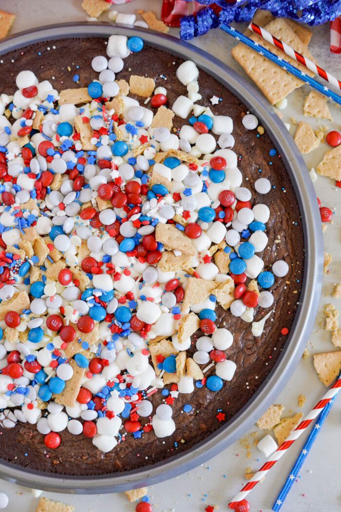 A round brownie topped with white icing, red, white, and blue candies, mini marshmallows, and graham cracker pieces on a table scattered with similar toppings and striped straws.