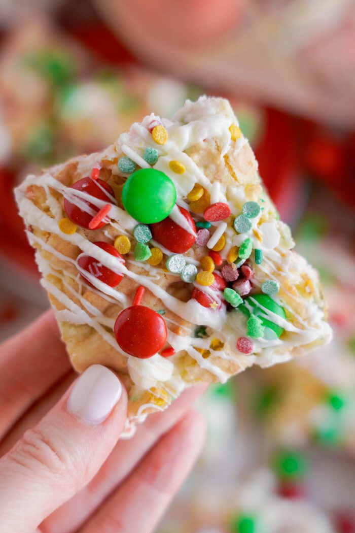 A close-up of a hand holding a Christmas Cookie Bar square covered in white icing, colorful sprinkles, and red and green candy-coated chocolates.